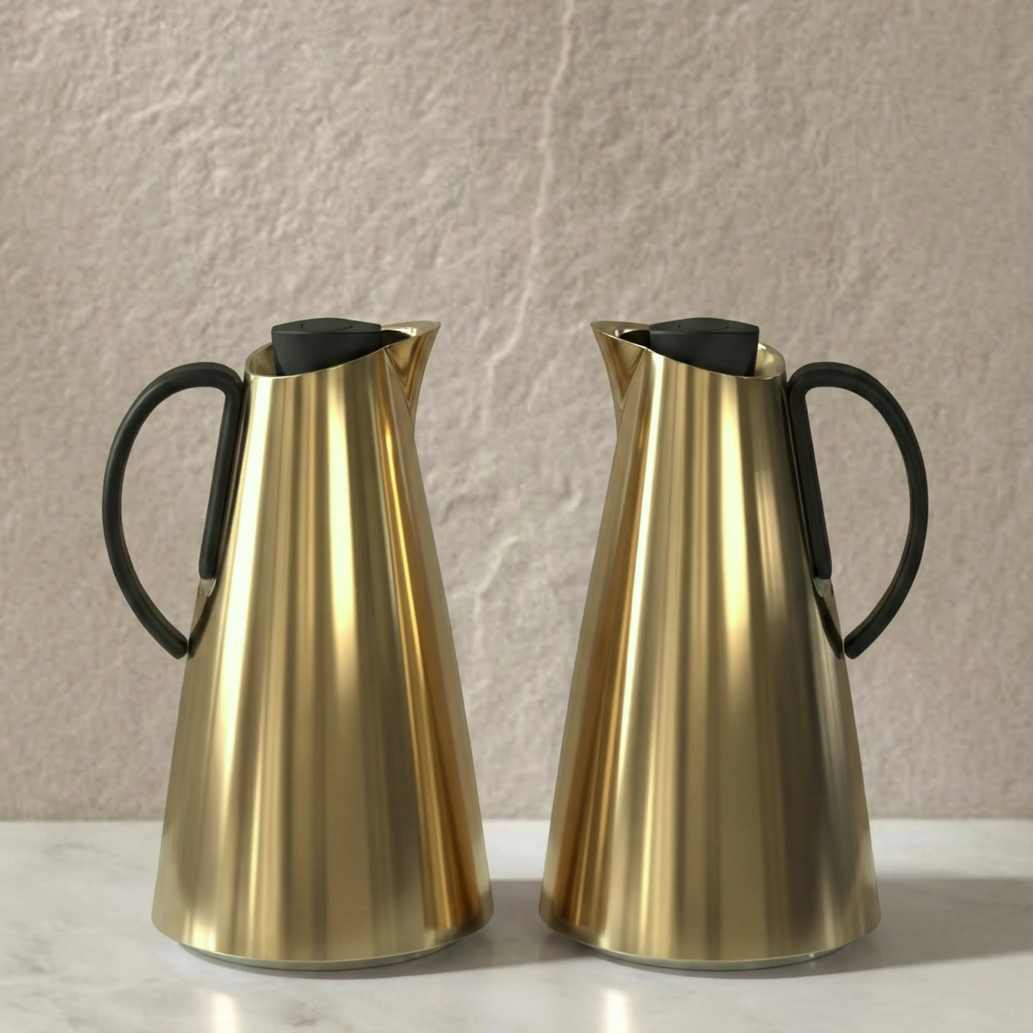 Two gold pitchers with black handles on a light surface against a beige wall.